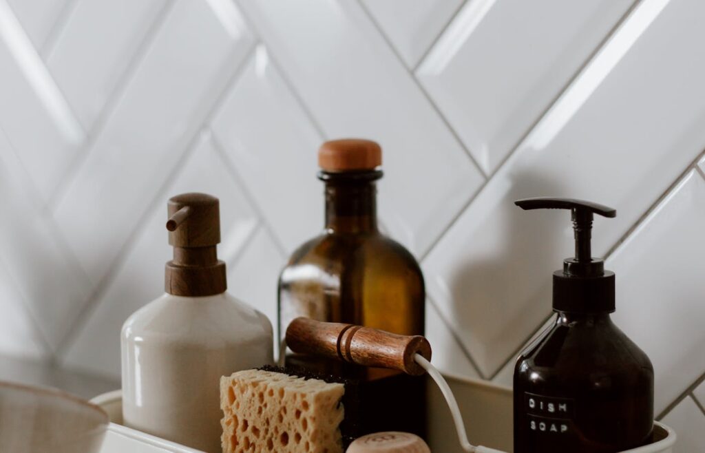 Cleaning product bottles and a sponge beside a tiled kitchen sink
